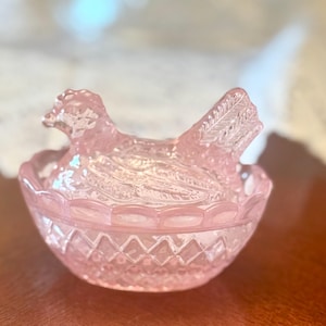 Könnte beinhalten: Eine durchscheinende, hellrosa Schale aus Pressglas mit einem Deckel in Form einer sitzenden Henne. Der Boden der Schale hat eine rautenförmige Textur, während der Deckel die detaillierte Form eines Vogels mit strukturierten Federn zeigt.