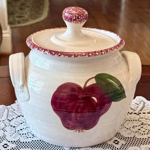 May include: White ceramic jar with a lid and handles. The jar features a large, red apple design with a green leaf. The lid and rim have a red speckled pattern. The jar is likely used for kitchen storage.