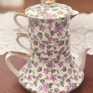 May include: A stackable tea set for one, featuring a white porcelain teapot, cup, and creamer decorated with a purple violet floral pattern and green leaves. Each piece has gold-trimmed handles and a gold knob on the lid, creating a vertical unit.