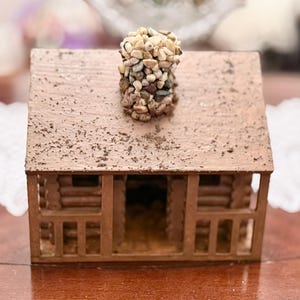 May include: A miniature brown wooden cabin with a stone chimney. The cabin features a log cabin design with a pitched roof and small porch. The roof is speckled with dark flecks, and the chimney is made of small, multi-colored stones.