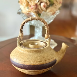 May include: A ceramic teapot with a bamboo handle. The teapot is tan with a dark brown band around the middle. The handle is made of curved bamboo pieces. The spout is short and curved. The teapot is sitting on a wooden surface.