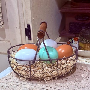 Puede incluir: Una cesta de alambre con un asa de madera llena de huevos de colores pastel. La cesta está sentada sobre un encaje blanco de ganchillo.