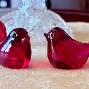 以下が含まれることがあります： 赤いガラス製の小さな鳥の置物2つ。鳥たちは向き合っており、光沢のある仕上げになっています。
