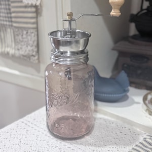 Peut inclure: Un moulin à café manuel de style vintage. Il comprend un bocal en verre transparent teinté de rose avec le mot "Ball" en relief. Le mécanisme du moulin est en acier inoxydable et possède une manivelle en bois. Le bocal est posé sur une nappe en dentelle blanche.