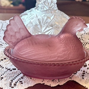 May include: A dusty rose-colored hen-shaped trinket box. The lid is shaped like a hen with detailed feather patterns. The base is a bowl-shaped nest with a beaded rim. The box is made of glass and sits on a white lace doily.