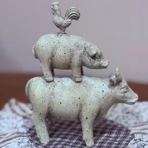 Puede incluir: Una figura decorativa de animales de granja apilados. Un gallo está encima de un cerdo, que está equilibrado sobre una vaca. Las figuras son de color gris claro moteado. La vaca tiene cuernos y el cerdo tiene un pequeño hocico. El gallo tiene una cresta detallada.