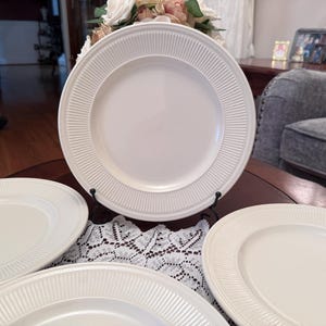 May include: A set of four off-white ceramic dinner plates featuring a smooth center and a wide, textured rim with vertical ribbed detailing. One plate is displayed upright on a black metal stand, while the other three plates are arranged below.