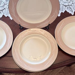 May include: A set of vintage dinner plates. The plates feature a pale pink border with a delicate white floral pattern and a gold rim. The plate centres are cream-coloured. The plates are stacked on top of each other.