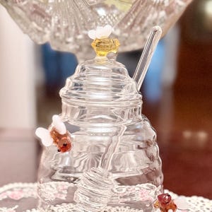 Puede incluir: Un tarro de miel de vidrio transparente con un cucharón de miel de vidrio. El tarro tiene una tapa decorativa con un acento de abeja amarilla y blanca. Dos acentos de abeja adicionales están unidos al tarro. El tarro está sobre un mantel de encaje.