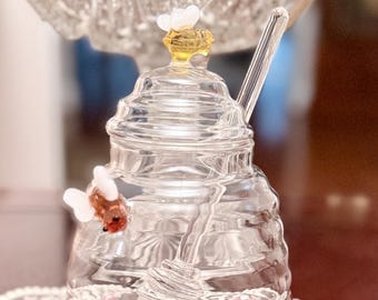 May include: A clear glass honey jar with a glass honey dipper. The jar has a decorative lid with a yellow and white bee accent. Two additional bee accents are attached to the jar. The jar is sitting on a lace tablecloth.