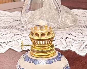 May include: A small oil lamp featuring a white ceramic base decorated with blue floral patterns. The lamp has a gold-colored metal burner assembly with an adjustment knob and a clear, ruffled-top glass chimney that sits atop the ceramic base.