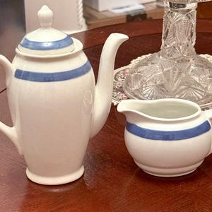 May include: White ceramic teapot and creamer set with blue stripes. The teapot has a lid and a curved spout. The creamer has a handle and a rounded shape. The set is on a wooden surface.