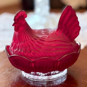 May include: A ruby red glass hen-shaped dish with a scalloped edge and gold trim. The hen is detailed with textured feathers and sits atop a clear, decorative base. This decorative piece is ideal for kitchen decor.