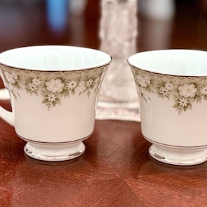 May include: Two white teacups with floral designs and gold trim. The cups have a pedestal base and curved handles. The floral pattern includes white flowers and green leaves. The cups are on a dark wooden surface.