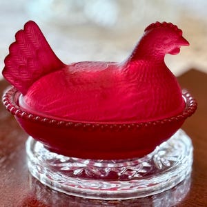 May include: A ruby red, vintage-style hen-shaped dish with a textured surface. The hen sits atop a matching base with a beaded rim. The dish is displayed on a clear, decorative glass coaster, showcasing a classic kitchenware design.