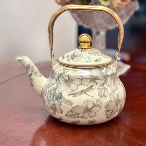 May include: Cream-colored teapot with a floral design in gray and white. The teapot has a gold-colored handle and lid finial. The floral pattern includes flowers and a dragonfly. A decorative item for the kitchen or dining room.