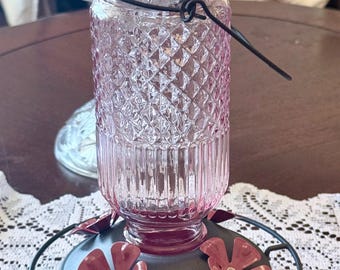 May include: A pink glass hummingbird feeder with a diamond pattern and a black metal base. The feeder has a circular base with two red flower-shaped feeding ports. A black wire handle is attached to the top.
