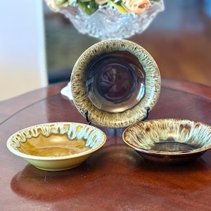 May include: Three ceramic bowls with a brown and tan glaze. The bowls are different sizes and shapes, with a glossy finish. The bowls are displayed on a dark wooden surface.