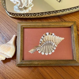 May include: A rectangular wooden frame containing a cross-stitch embroidery of a tan and brown seashell centered on a solid terracotta-colored fabric background. The embroidery features detailed shading and white highlights on the shell's ridges.