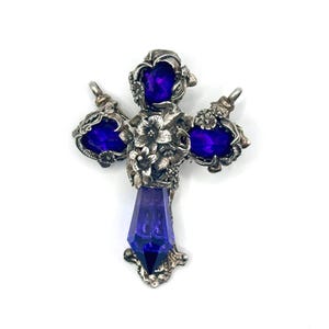 May include: A silver cross pendant with a purple crystal in the center and three smaller purple crystals on the arms. The cross is decorated with floral details.