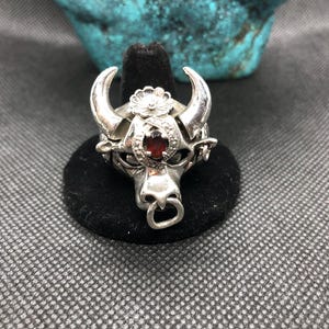 May include: A silver ring featuring a bull's head design. The bull's head has a red gemstone set in the centre of its forehead and a silver ring around its nose.