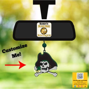 May include: A car air freshener hanging from a rearview mirror, featuring a black pirate hat and skull design with crossbones. The text ROBIN'S BUSY BEE BOUTIQUE is visible on the mirror, with the text Customize Me! and a red arrow pointing.