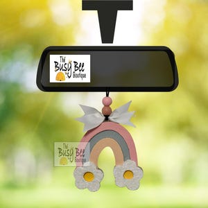 May include: A car rearview mirror hanging ornament featuring a rainbow shape with a white bow and two wooden beads. The rainbow has pink, grey, and peach arches with white flower accents at the base. The text The Busy Bee Boutique is visible.
