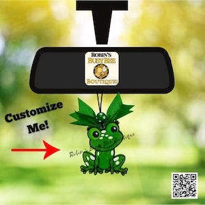 May include: A green frog ornament hanging from a black rearview mirror. The frog features white spots, large black eyes, and a green ribbon bow on top. The text "ROBIN'S BUSY BEE BOUTIQUE" is visible on the mirror, with "Customize Me!" and an arrow.
