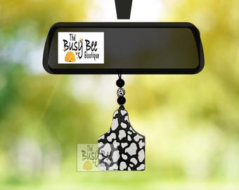 May include: A car rearview mirror accessory featuring a black and white cow print tag suspended by a black cord with black and white beads. The tag displays the text The Busy Bee Boutique with a small yellow beehive icon graphic.