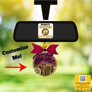 May include: A car air freshener with a burgundy bow and a round pendant that says "Be the Sunshine". The air freshener hangs from a car's rearview mirror. The text "Customize Me!" is also visible.