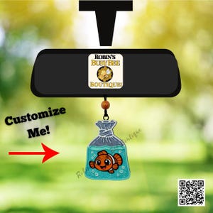 May include: A car rearview mirror charm featuring a rectangular black base with a Robin's Busy Bee Boutique logo. A dangling acrylic pendant shows a cartoon orange clownfish inside a teal glittery plastic bag, suspended by a brown bead and cord.