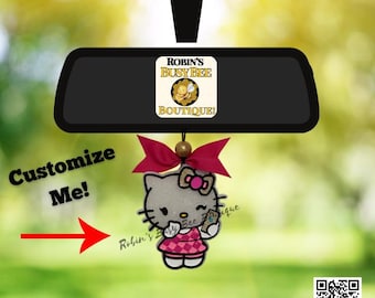 May include: A car rearview mirror hanging ornament featuring a glittery white cat character wearing a pink plaid dress and a pink bow. The ornament is suspended by a string with a gold bead. Text reads Robin's Busy Bee Boutique and Customize Me.