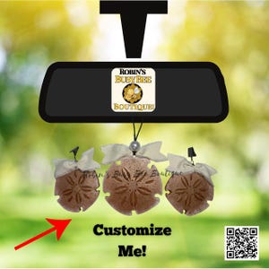 May include: A black car rearview mirror accessory featuring a white square logo with the text Robin's Busy Bee Boutique and a bee graphic. Three tan sand dollar ornaments with white ribbon bows hang from the mirror by black strings.