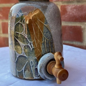 May include: A ceramic water dispenser with a cork stopper and a wooden spigot. The jar has a mottled grey and brown glaze with leaf-like patterns in green and gold. The spigot is made of light-coloured wood.