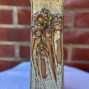 May include: A rectangular ceramic vase with a textured surface and a beige, brown, and tan colour scheme. The vase features raised circular and linear designs. The top of the vase has a slightly flared opening.