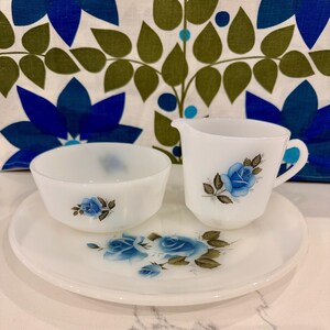 May include: A white milk glass dish set with a blue rose floral design. The set includes a small bowl, a creamer with a handle, and a round plate. The background features a blue and green floral pattern.