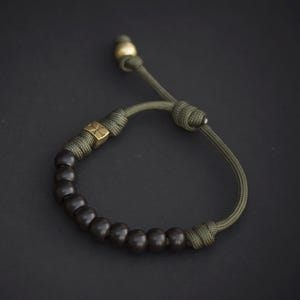 May include: A bracelet featuring a series of dark brown spherical beads and a single square brass bead with an etched cross design, all strung on olive green paracord with an adjustable sliding knot closure and a small brass bead at the end.