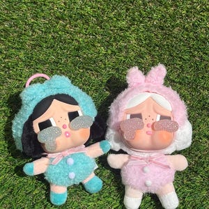Puede incluir: Dos juguetes de peluche con gafas de sol brillantes. Un juguete es azul claro con cabello negro y el otro es rosa con orejas de conejo blancas.