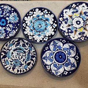 May include: Five round ceramic plates with intricate blue and white floral designs. Each plate features a unique pattern, with a dark blue border and a light blue and white central motif. The plates are arranged on a neutral surface.