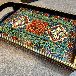 Puede incluir: Bandeja de servicio rectangular de madera negra con asa. Decorada con un colorido patrón geométrico en rojo, azul, amarillo y blanco. El diseño presenta formas florales y geométricas.