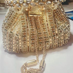 May include: A gold beaded evening bag with a drawstring closure and a decorative tassel. The bag features intricate beadwork and a jeweled handle. The bag is a light gold colour and is designed for formal occasions.