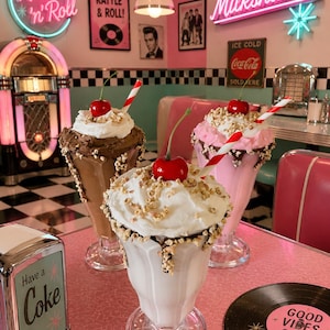 May include: Three milkshakes in glass pedestal cups topped with whipped cream, chopped nuts, and a bright red cherry. One milkshake is chocolate, one is strawberry, and one is vanilla, each featuring a red and white striped paper straw.