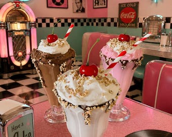 Può includere: Tre bicchieri da milkshake riempiti con gelato al cioccolato, fragola e vaniglia, guarniti con panna montata, granella di noci, una ciliegia rossa e una cannuccia a righe rosse e bianche, su un tavolo rosa con portatovaglioli Coke e vinile.
