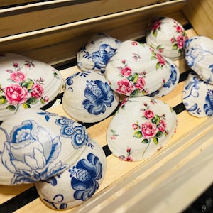 May include: Decorative seashells with floral and blue patterns. The shells are white with pink roses and green leaves, and blue floral designs. They are arranged in a wooden crate.