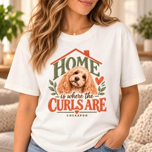 Può includere: Una t-shirt bianca con la grafica di un cane Cockapoo marrone. Il testo "HOME is where the CURLS ARE" è scritto in verde e arancione. Sopra il cane c'è il contorno rosso di una casa, con foglie verdi e un piccolo cuore rosso come decoro.