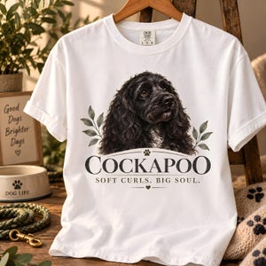 Könnte beinhalten: Ein weißes kurzärmeliges T-Shirt mit einer realistischen Illustration eines schwarzen Cockapoo-Hundes mit lockigem Fell. Unter dem Hund steht der Text COCKAPOO und SOFT CURLS. BIG SOUL. mit einer kleinen Pfotenabdruck- und Herzgrafik.