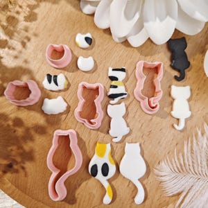 Puede incluir: Una colección de cortadores de galletas con forma de gato y decoraciones de fondant con forma de gato. Los cortadores son rosas y las decoraciones de fondant son blancas, negras, amarillas y marrones. Los artículos están dispuestos sobre una superficie de madera.