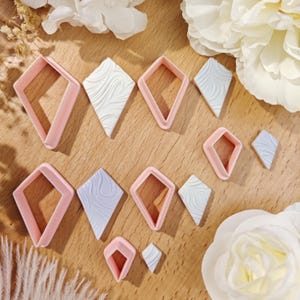 May include: A collection of pink diamond-shaped cookie cutters and diamond-shaped clay pieces with a marbled design in white and light blue. The cutters are in various sizes, and the clay pieces are arranged on a wooden surface.