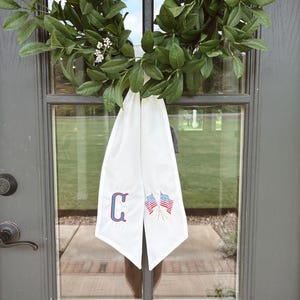 May include: A green wreath hangs on a gray door, with a white fabric banner. The banner features a blue embroidered letter "C" and two small American flags. The wreath is made of green leaves and small white flowers.