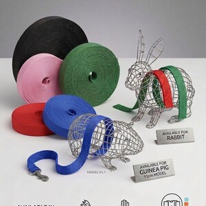 May include: Multiple rolls of nylon webbing in black, pink, green, red, and blue. Two wire animal sculptures, a rabbit and a guinea pig, are displayed with webbing straps. Small signs read AVAILABLE FOR RABBIT and AVAILABLE FOR GUINEA PIG 11cm MODEL.
